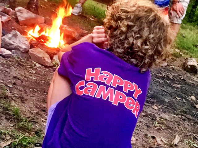 child sitting at the campfire with a happy camper shirt on.